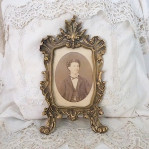 May include: An antique gold-coloured picture frame with an oval portrait of a man in a suit and bow tie. The frame has a detailed, decorative design and is propped up on a stand. The frame is set against a white lace background.