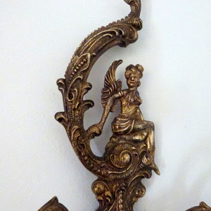 K&ouml;nnte beinhalten: Verzierter Wandhaken aus goldenem Metall mit einem dekorativen Schn&ouml;rkeldesign und einer Cherubfigur. Der Haken hat drei Arme zum Aufh&auml;ngen von Gegenst&auml;nden.