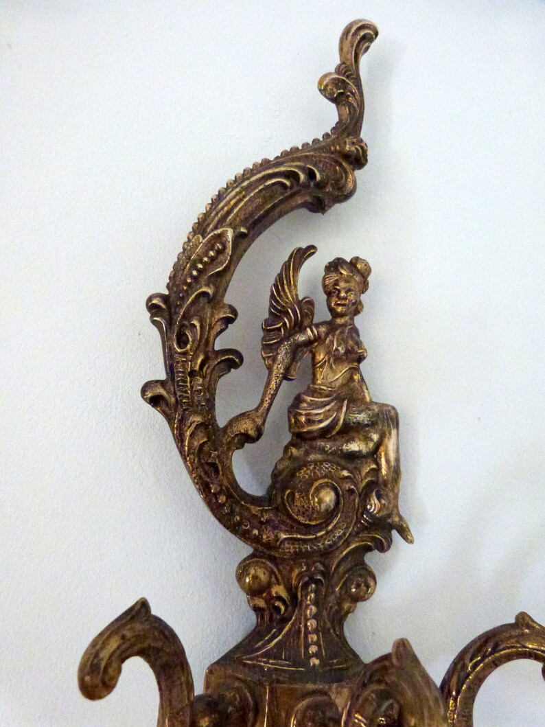 K&ouml;nnte beinhalten: Verzierter Wandhaken aus goldenem Metall mit einem dekorativen Schn&ouml;rkeldesign und einer Cherubfigur. Der Haken hat drei Arme zum Aufh&auml;ngen von Gegenst&auml;nden.