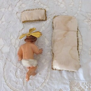 May include: A vintage figurine of a baby Jesus with a gold halo, a small pillow, and a larger pillow. The baby is nude, with a white diaper. The pillows are cream-colored with gold trim. The items are on a white, embroidered surface.