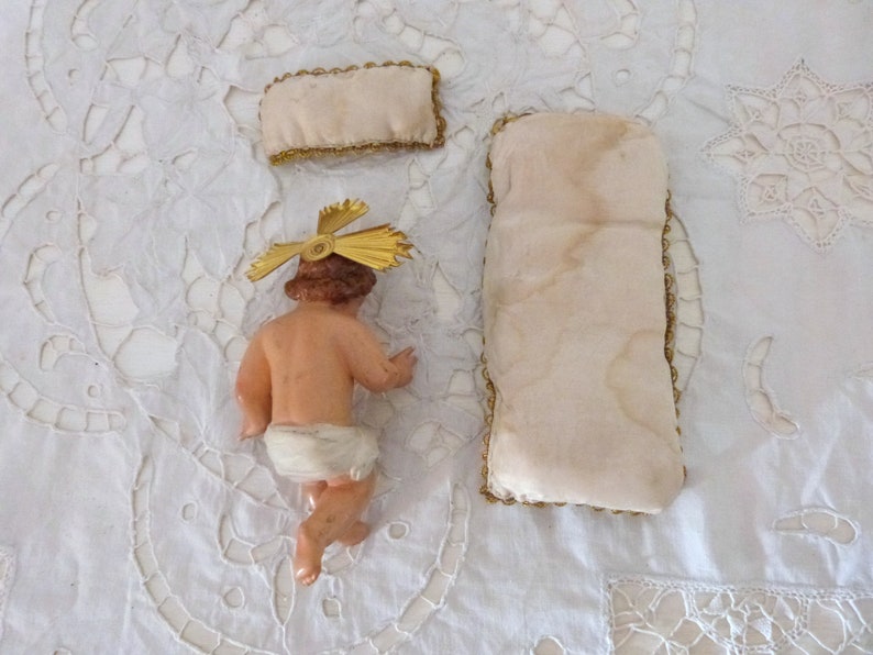 May include: A vintage figurine of a baby Jesus with a gold halo, a small pillow, and a larger pillow. The baby is nude, with a white diaper. The pillows are cream-colored with gold trim. The items are on a white, embroidered surface.
