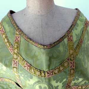 May include: Close-up of a green liturgical vestment with a floral pattern. The vestment has decorative gold and burgundy trim around the neckline and along the seams. The fabric is a light green color.