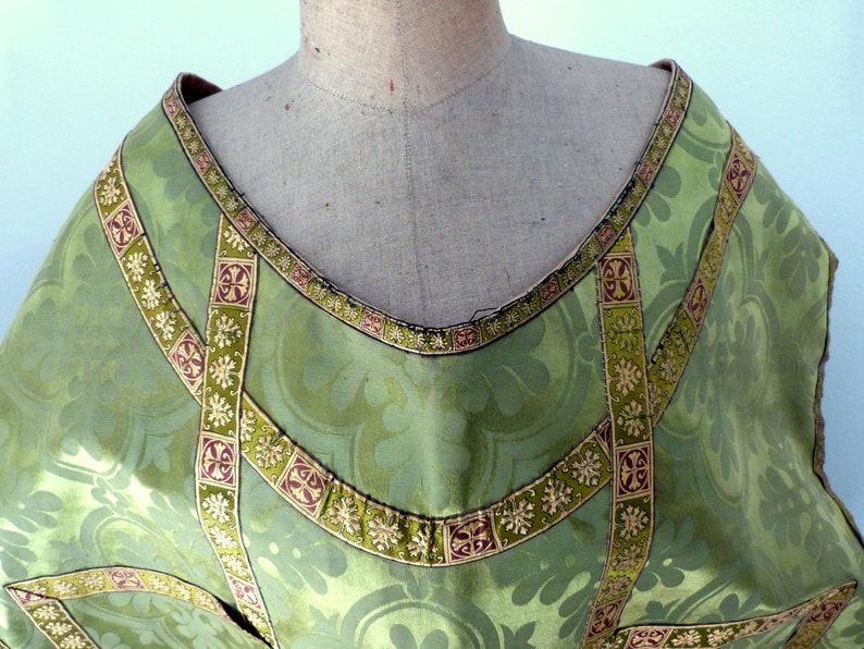 May include: Close-up of a green liturgical vestment with a floral pattern. The vestment has decorative gold and burgundy trim around the neckline and along the seams. The fabric is a light green color.
