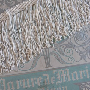 May include: White fringe trim with a long, thin texture. The fringe is made of small, tightly woven strands of yarn.