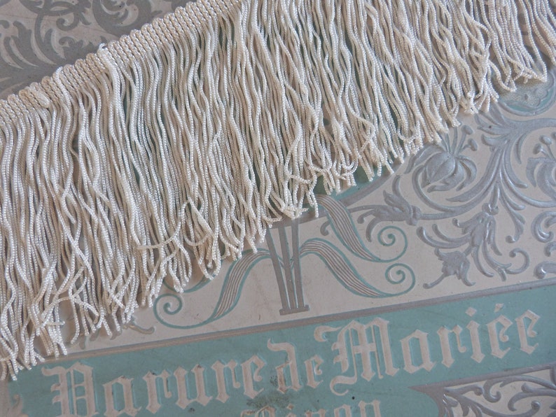 May include: White fringe trim with a long, thin texture. The fringe is made of small, tightly woven strands of yarn.