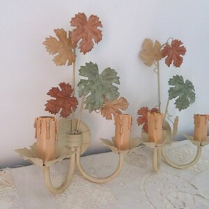 May include: A pair of vintage wall sconces with a grapevine design. The sconces are painted in a cream color and feature faux candles with a light peach color. The leaves are painted in shades of green, brown, and peach.