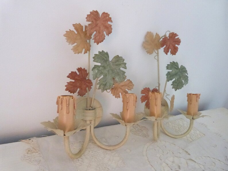 May include: A pair of vintage wall sconces with a grapevine design. The sconces are painted in a cream color and feature faux candles with a light peach color. The leaves are painted in shades of green, brown, and peach.