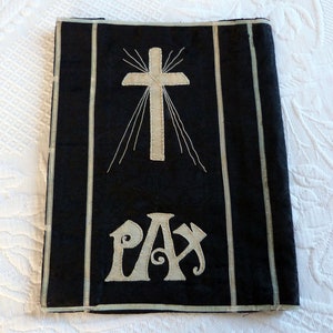 Op de afbeelding: Een zwarte stoffen boekomslag met een wit geborduurd kruis en het woord "PAX" in een decoratief lettertype.