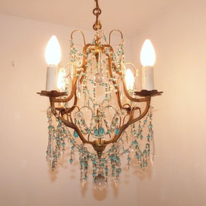 May include: An antique chandelier with a bronze-colored frame, adorned with clear and turquoise-colored crystal beads and teardrop pendants. The chandelier has four candle-style light fixtures with lit bulbs, creating a warm glow.