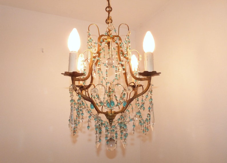 May include: An antique chandelier with a bronze-colored frame, adorned with clear and turquoise-colored crystal beads and teardrop pendants. The chandelier has four candle-style light fixtures with lit bulbs, creating a warm glow.
