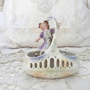 Op de afbeelding: Een vintage porseleinen mand met een handvat, met een beeldje van een meisje in een paarse jurk. De mand heeft een opengewerkt ontwerp en is geschilderd in pastelkleuren, met gouden accenten. De mand staat op een wit kanten tafelkleed.