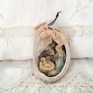 Può includere: Ornamento natalizio ovale con una scena della Natività. L'ornamento ha una cornice color crema con un fiocco rosa e un cordino nero per appenderlo. Lo sfondo è un tessuto bianco ricamato.