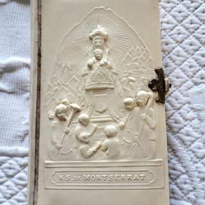 May include: Ivory-coloured religious book with a detailed relief carving on the cover. The carving depicts a religious figure with a halo, surrounded by figures and the text "N.S. de Montserrat". A metal clasp secures the book.