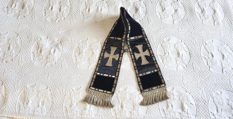 May include: A dark blue scarf with a gold cross design and fringe. The scarf has a velvet texture and is decorated with gold trim. The crosses are centered on the scarf.