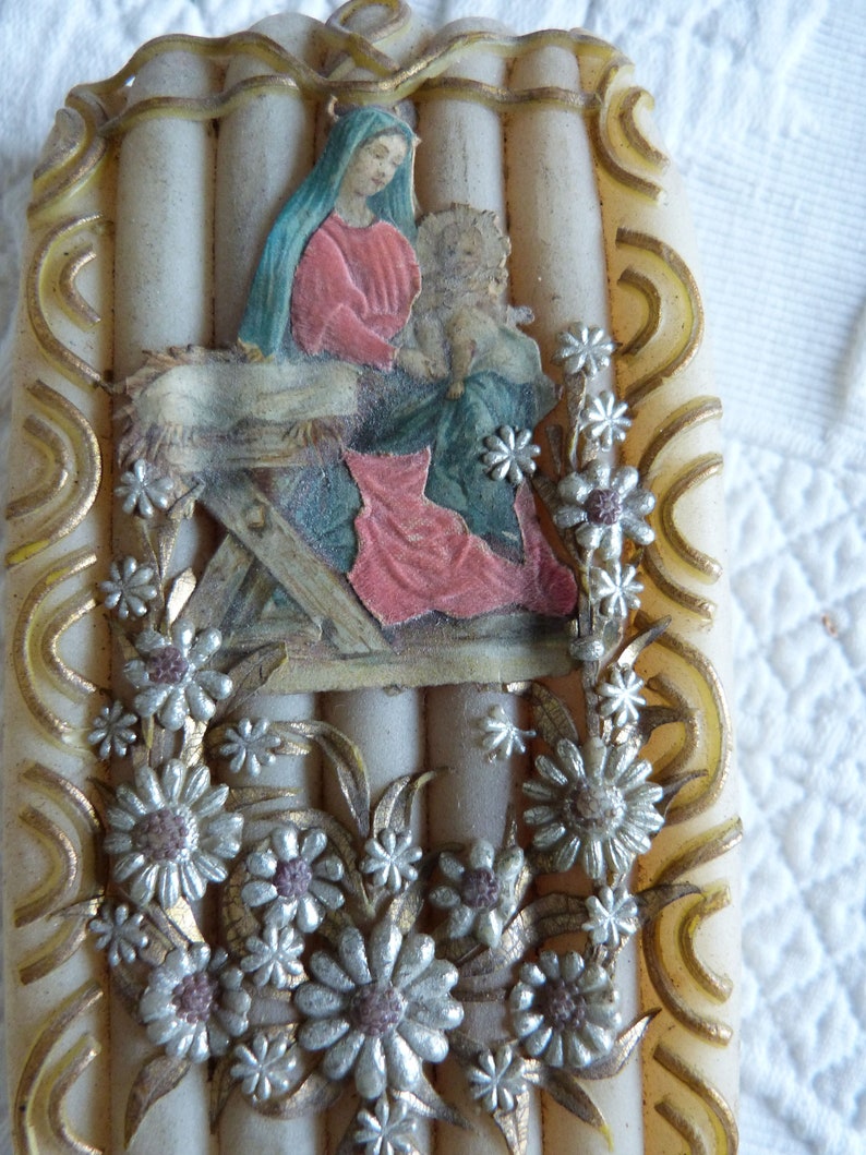May include: A white candle with a religious image of Mary and baby Jesus in a manger. The image is surrounded by silver and gold floral accents.