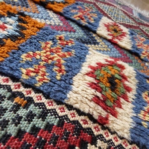 Op de afbeelding: Een kleurrijk Marokkaans tapijt met ingewikkelde geometrische patronen in tinten blauw, rood, oranje, groen en wit. Het tapijt is geweven met een dikke, textuurrijke pool.