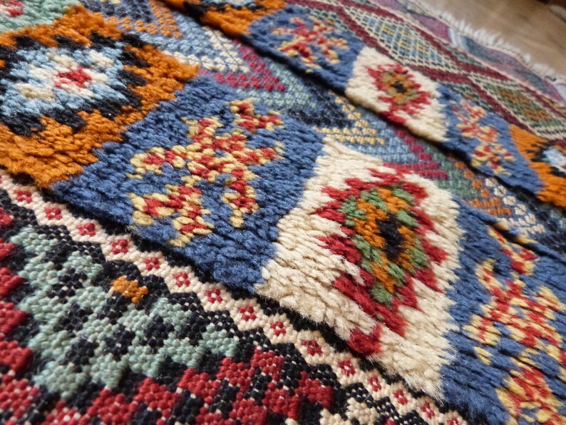 Op de afbeelding: Een kleurrijk Marokkaans tapijt met ingewikkelde geometrische patronen in tinten blauw, rood, oranje, groen en wit. Het tapijt is geweven met een dikke, textuurrijke pool.