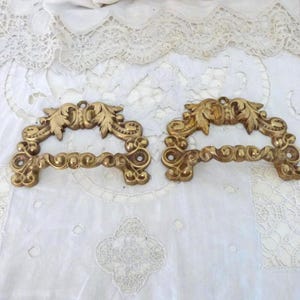 Op de afbeelding: Twee antieke messing lade handgrepen met sierlijke, bloemmotieven. Elke handgreep heeft een gebogen handvat met kleine, decoratieve bollen en een bovenste gedeelte met gestileerde bladeren. Geplaatst op een wit, met kant bedekt oppervlak.