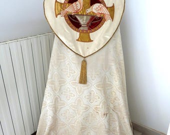 May include: Cream-colored religious vestment with a gold cross design on the back. The cross features two doves and a chalice. The vestment has a gold tassel and a patterned fabric.