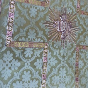 May include: Close-up of a green fabric with a repeating floral pattern. The fabric is trimmed with decorative borders in gold and purple. A central embroidered design features the letters "IHS" within a sunburst.