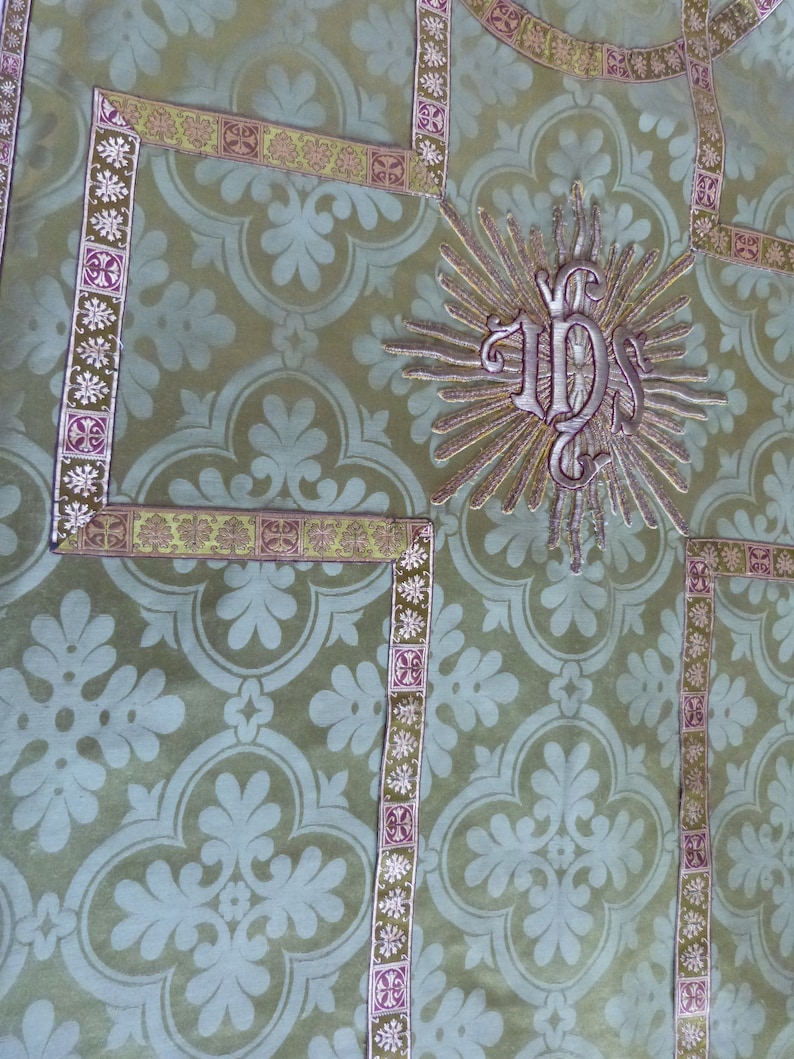 May include: Close-up of a green fabric with a repeating floral pattern. The fabric is trimmed with decorative borders in gold and purple. A central embroidered design features the letters "IHS" within a sunburst.