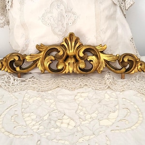 Può includere: Un elemento architettonico decorativo dorato e ornato. Il pezzo presenta un design simmetrico con dettagli a volute e un motivo floreale centrale stilizzato. È probabilmente in metallo e ha un'estetica vintage o antica. Il pezzo è lungo circa 46 cm.