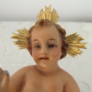 May include: A vintage figurine of a baby with a halo. The baby has light skin, rosy cheeks, and brown hair. A gold-colored halo is behind the baby's head. The figurine is likely made of porcelain or a similar material.
