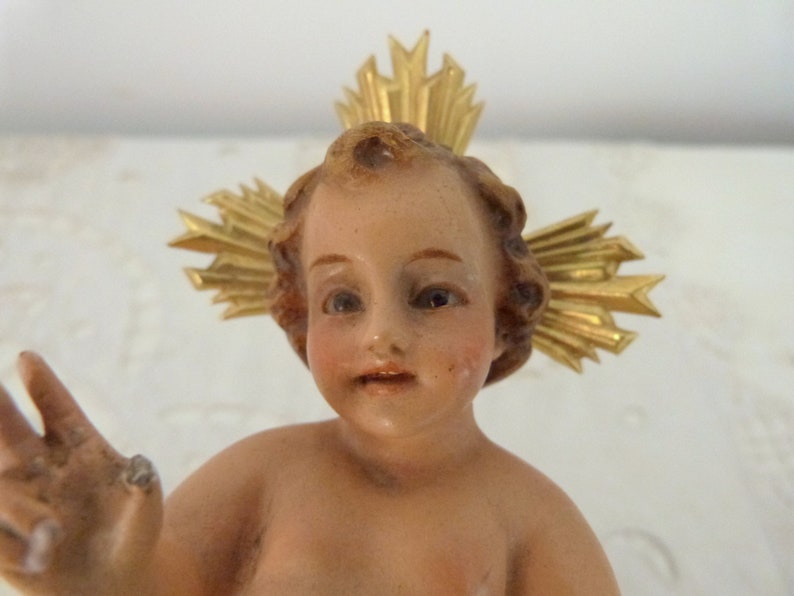May include: A vintage figurine of a baby with a halo. The baby has light skin, rosy cheeks, and brown hair. A gold-colored halo is behind the baby's head. The figurine is likely made of porcelain or a similar material.