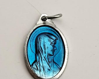 May include: An oval-shaped metal pendant featuring a profile relief of a figure wearing a head covering, set against a vibrant blue enamel background. The pendant is framed in a silver-toned metal border and includes a small loop at the top.