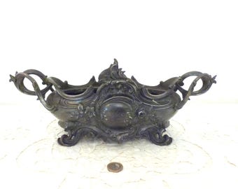 Op de afbeelding: Een antieke zilverkleurige centerpiece kom met sierlijke krullen en bloemendetails. De kom heeft twee handvatten en rust op decoratieve voetjes. Een kleine munt is zichtbaar op de voorgrond.
