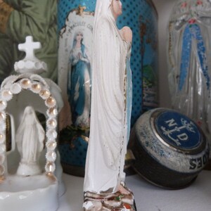 May include: A white porcelain figurine of the Virgin Mary, standing on a brown base with green and white accents. The figurine is wearing a white robe and a blue sash. The figurine is surrounded by other decorative items, including a blue and white tin can with a religious image and a blue and white glass bottle.