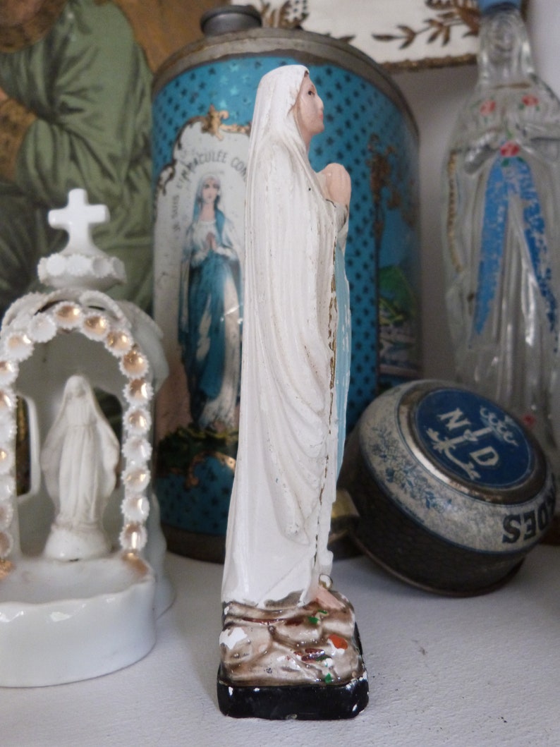 May include: A white porcelain figurine of the Virgin Mary, standing on a brown base with green and white accents. The figurine is wearing a white robe and a blue sash. The figurine is surrounded by other decorative items, including a blue and white tin can with a religious image and a blue and white glass bottle.