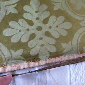 May include: Close-up of a section of fabric with a repeating floral pattern in cream against a green background. The fabric has a decorative border with a cream-colored backing and a white surface.