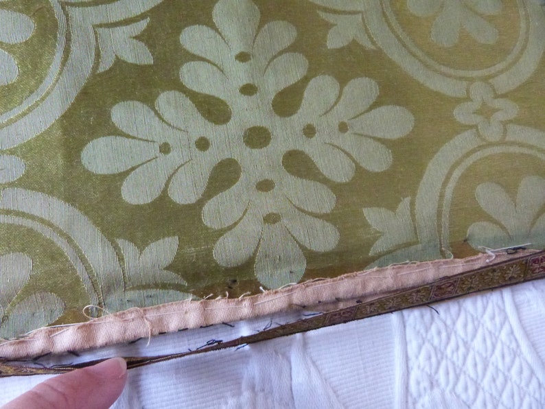 May include: Close-up of a section of fabric with a repeating floral pattern in cream against a green background. The fabric has a decorative border with a cream-colored backing and a white surface.