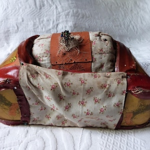 Pode incluir: Um almofadinha para alfinetes vintage com um exterior de tecido vermelho, marrom e branco. O almofadinha é preenchido com um tecido branco e tem um grupo de alfinetes no topo. O almofadinha está sobre uma superfície branca.