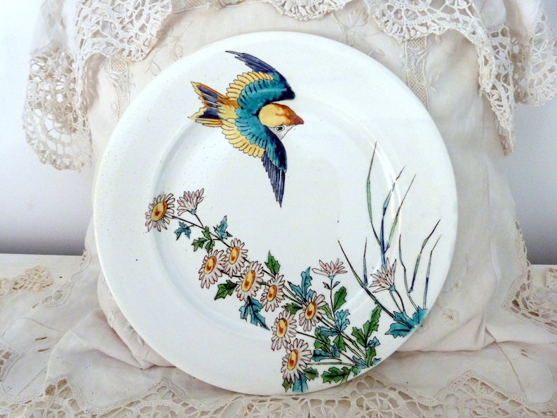 Pode incluir: Um prato de cer&acirc;mica branco com um desenho de p&aacute;ssaro azul e amarelo. O p&aacute;ssaro est&aacute; voando sobre um grupo de flores brancas com folhas verdes. O prato tem um fundo branco com uma textura sutilmente salpicada.