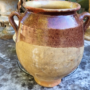 Peut inclure: Pot en céramique rustique à deux anses et à large ouverture. Le pot présente des bandes horizontales de couleur : une base beige clair, une section centrale marron texturée et un bord marron plus foncé. Le pot est probablement vintage ou antique.