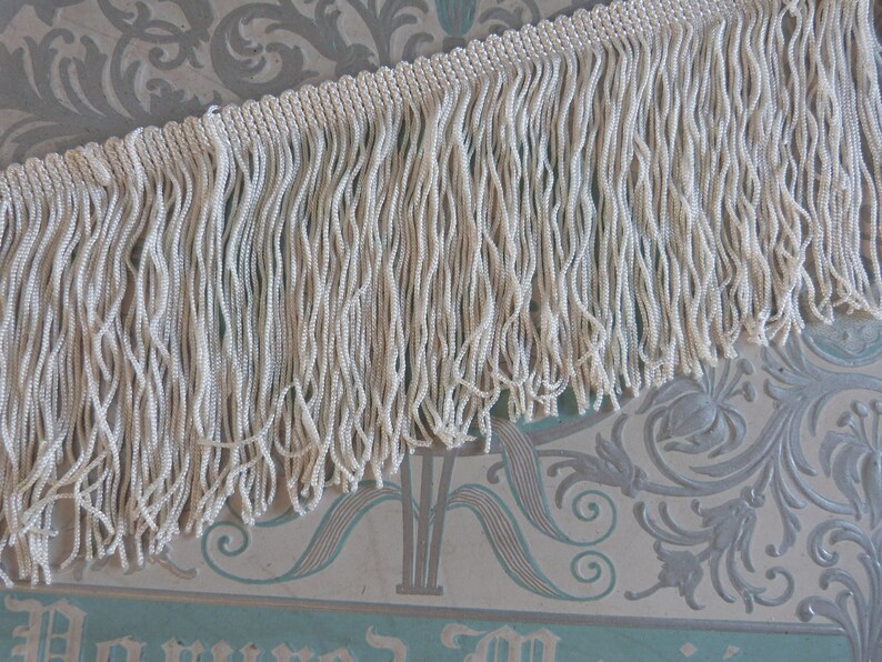 May include: White fringe trim with a beaded texture. The fringe is approximately 1 inch in length.