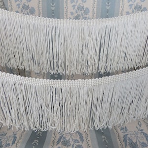 May include: Two white cotton fringe trims with a woven edge. The fringe is approximately 2 inches long.