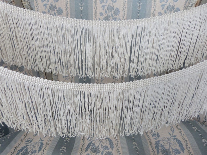 May include: Two white cotton fringe trims with a woven edge. The fringe is approximately 2 inches long.