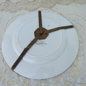 May include: The back of a white ceramic plate with a brass plate hanger attached. The plate has a printed design on the back.