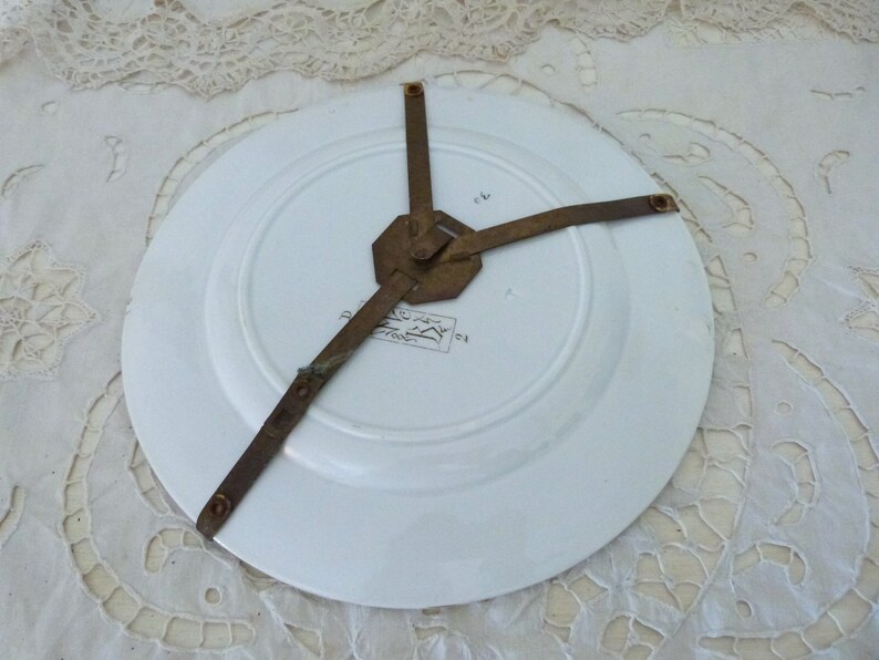 May include: The back of a white ceramic plate with a brass plate hanger attached. The plate has a printed design on the back.