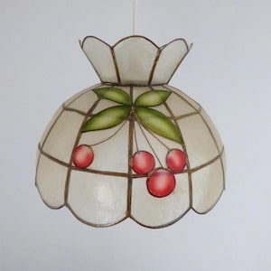 Puede incluir: Una lámpara colgante decorativa con una parte superior blanca y festoneada y una pantalla redondeada. La pantalla presenta un patrón de cuadrícula con cerezas rojas pintadas y hojas verdes. La lámpara cuelga de un cable blanco.