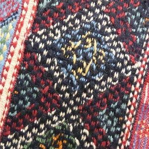 Op de afbeelding: Close-up van een kleurrijk geweven stof met een geometrisch patroon. De stof is gemaakt van draden in verschillende kleuren, waaronder rood, blauw, wit en zwart. Het patroon is ingewikkeld en gedetailleerd, en de stof heeft een textuur.