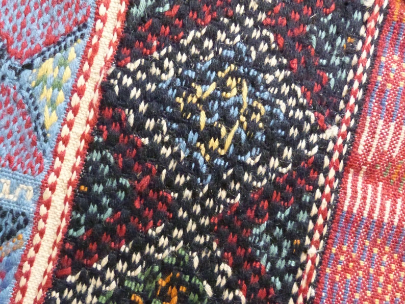 Op de afbeelding: Close-up van een kleurrijk geweven stof met een geometrisch patroon. De stof is gemaakt van draden in verschillende kleuren, waaronder rood, blauw, wit en zwart. Het patroon is ingewikkeld en gedetailleerd, en de stof heeft een textuur.