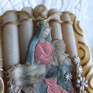 May include: A vintage religious image of the Virgin Mary holding baby Jesus. The image is framed in a white and gold decorative frame with a floral design. The image is surrounded by white candles.