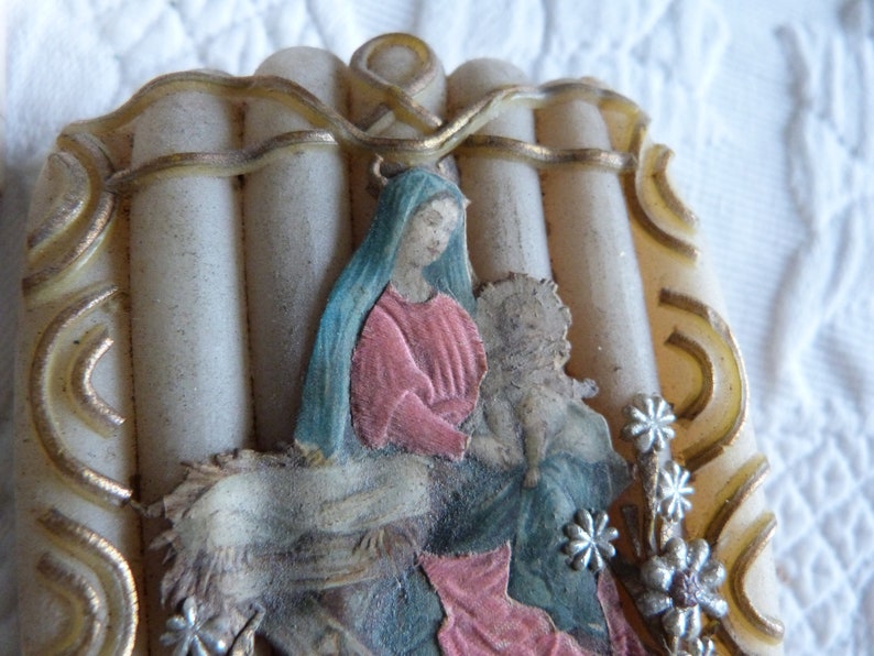 May include: A vintage religious image of the Virgin Mary holding baby Jesus. The image is framed in a white and gold decorative frame with a floral design. The image is surrounded by white candles.
