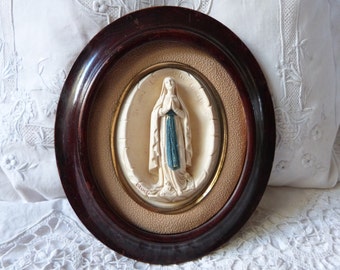 May include: An oval framed religious image of the Virgin Mary in a white dress and blue robe. The image is in a cream-colored oval frame with a gold inner border. The frame is dark brown wood.