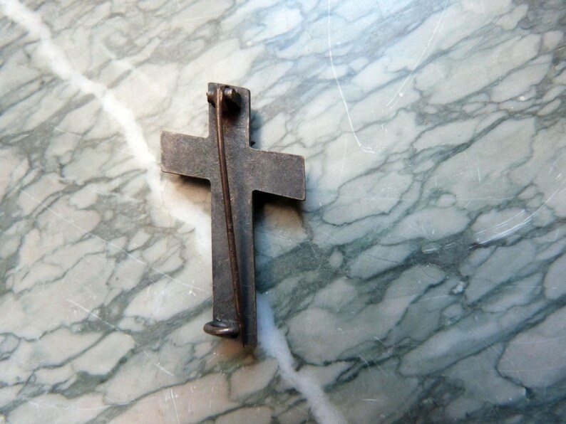 May include: A silver-toned cross pin with a pin back. The cross is slightly raised and has a textured surface.