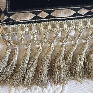 May include: Close-up of a decorative textile with a black velvet panel and a geometric pattern in gold and beige. The bottom edge features a fringe of gold tassels, adding a touch of elegance. The textile is likely used for home decor.